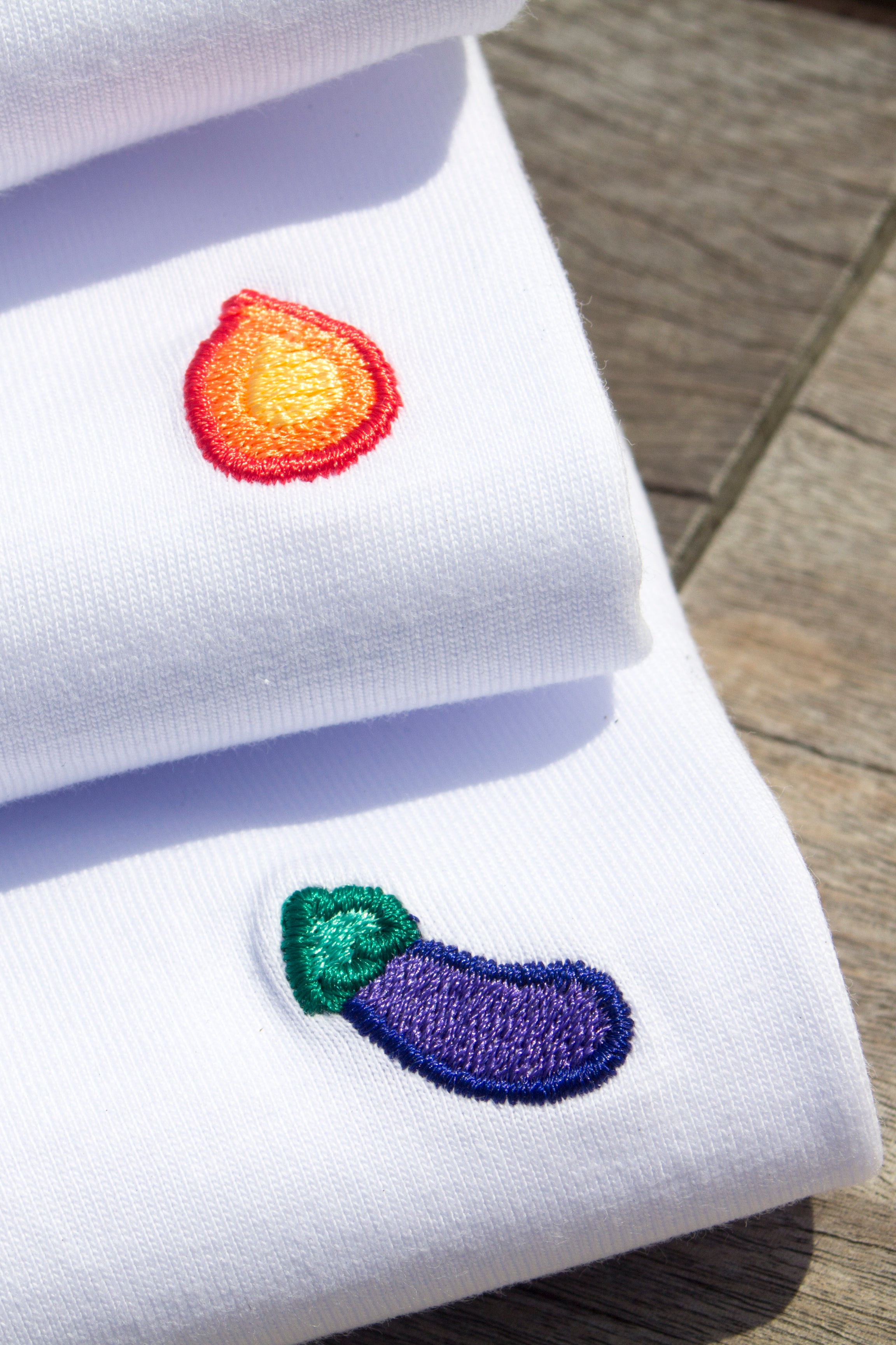 Close-up of flame and eggplant embroidery details on stacked white Out Now! t-shirts, showcasing the intricate stitching and bold designs inspired by LGBTQ+ pride.